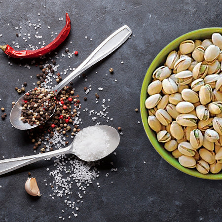 Spices, including red pepper and black peppercorns, on a dark surface with a bowl of beans.
